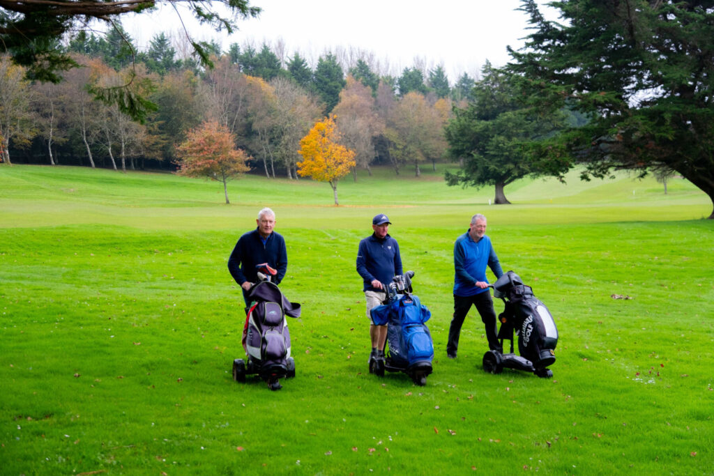 Three men with golf bags walking on a lush green golf course, surrounded by autumn trees.