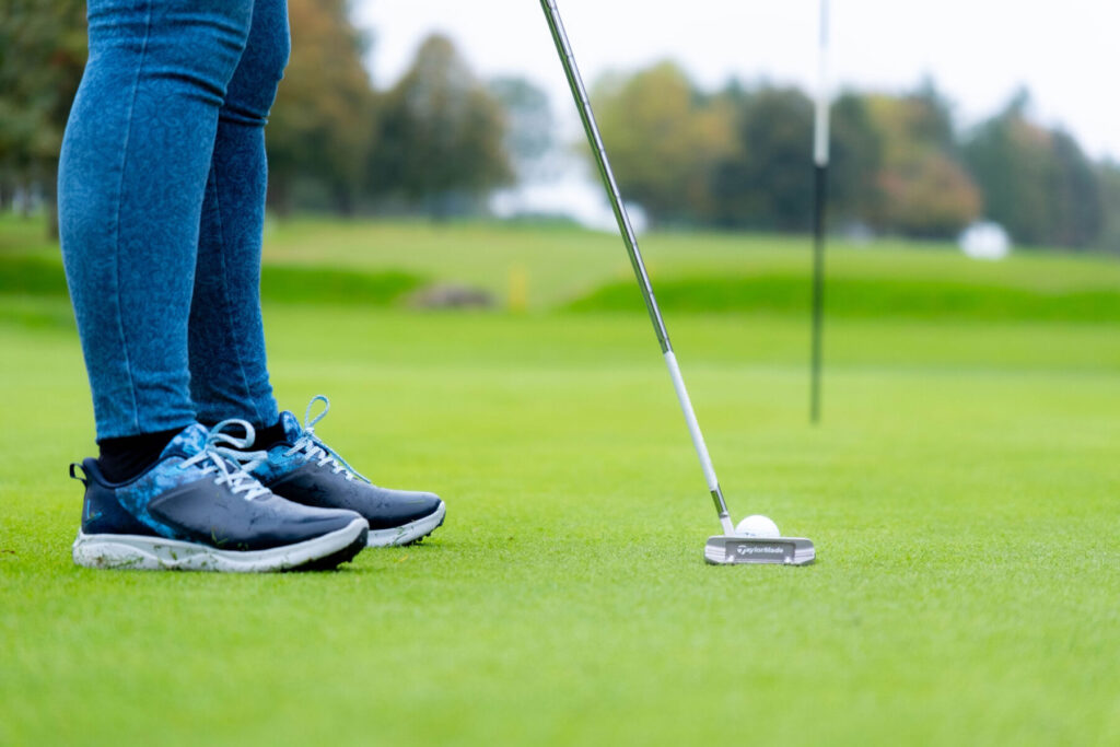 Golfer putting on a green with a TaylorMade putter, wearing blue jeans and sneakers.