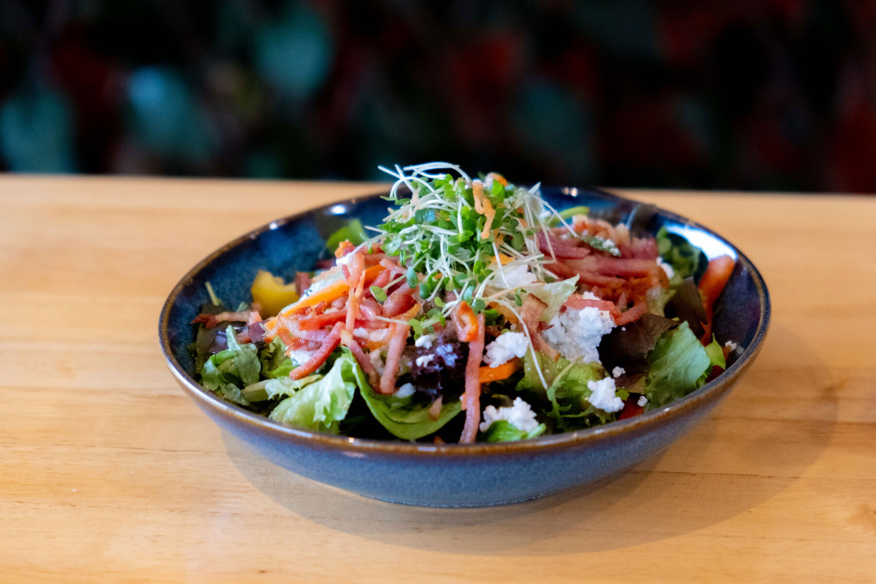 Fresh salad with mixed greens, shredded carrots, crumbled cheese, and sprouts in a blue bowl on a wooden table.