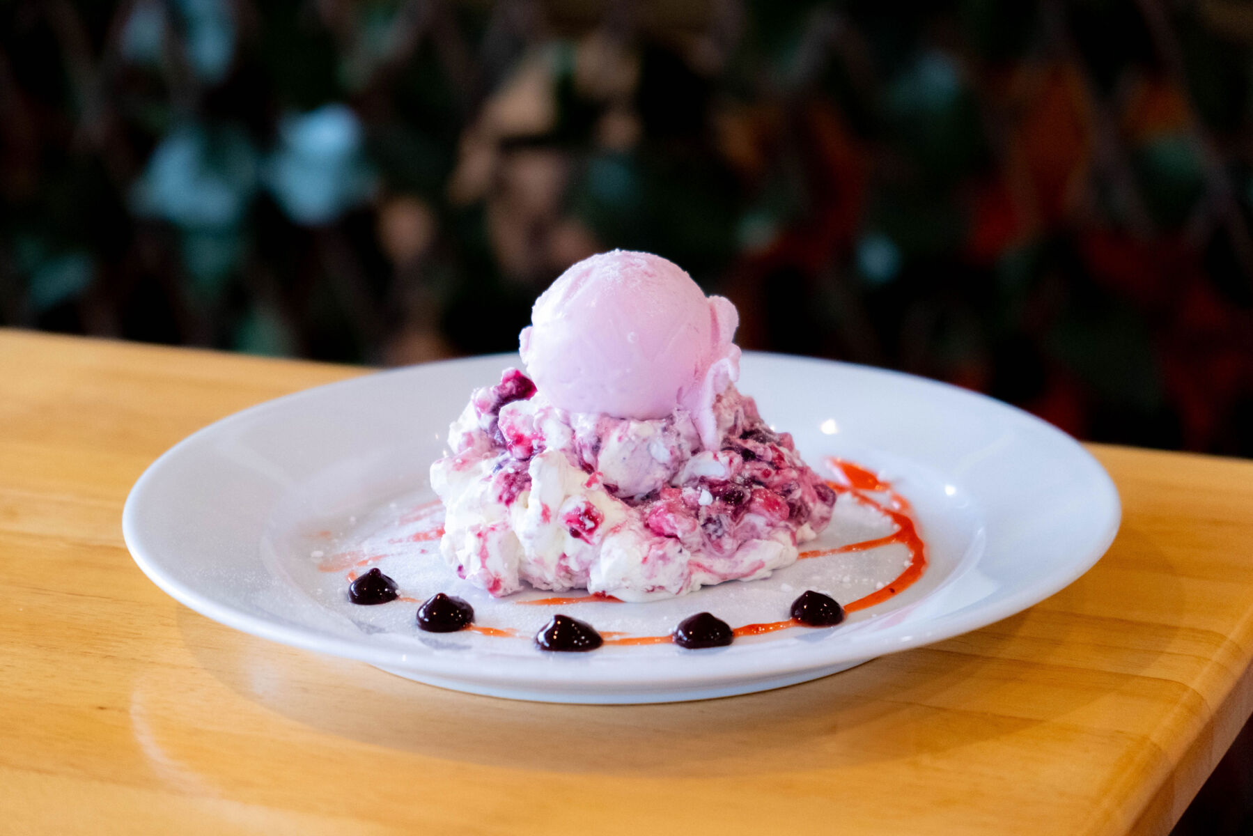 Berry pavlova topped with pink ice cream on a plate with fruit drizzle and garnish.
