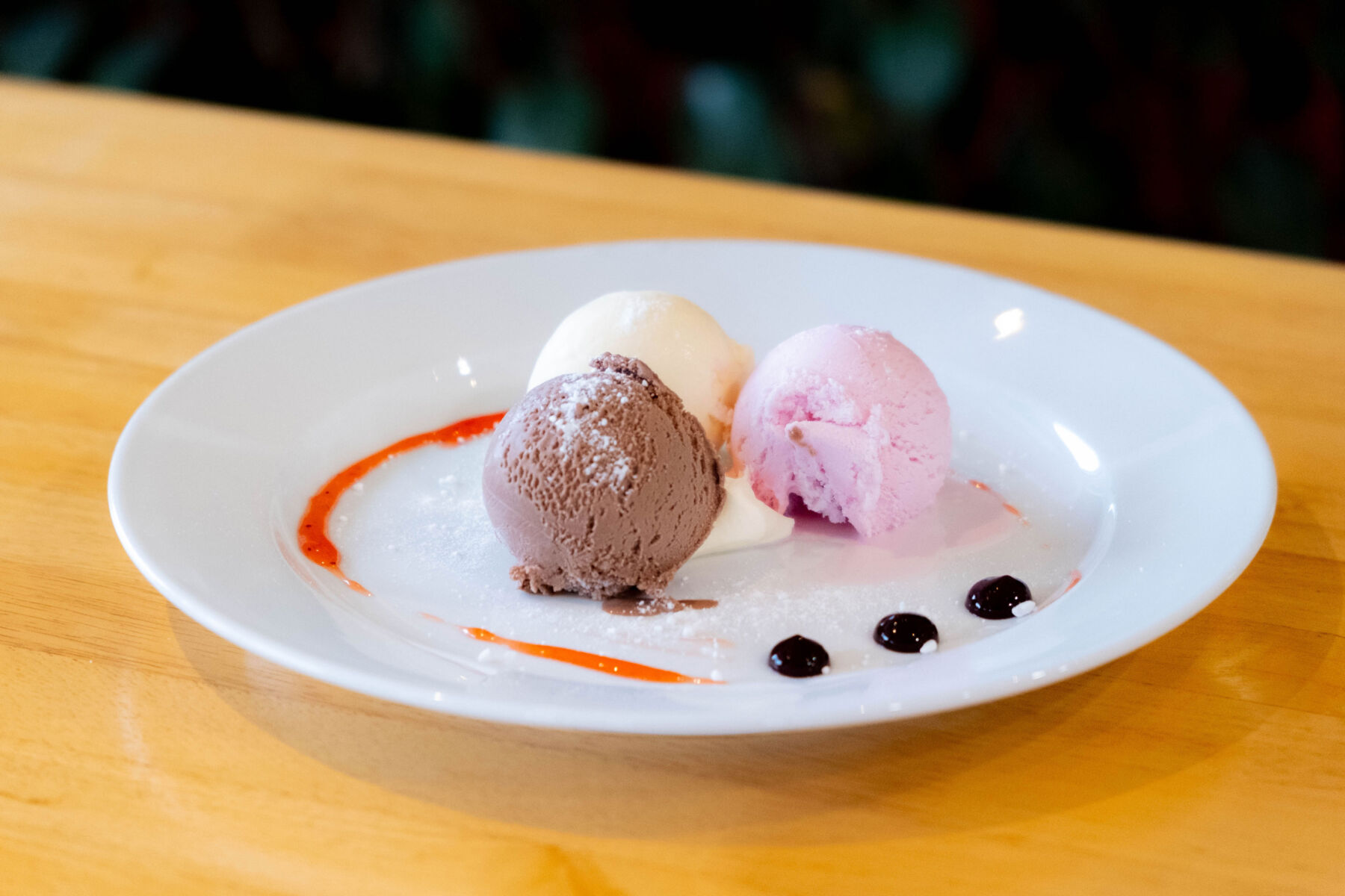 Three scoops of ice cream (chocolate, vanilla, strawberry) with red and dark syrup on a white plate.