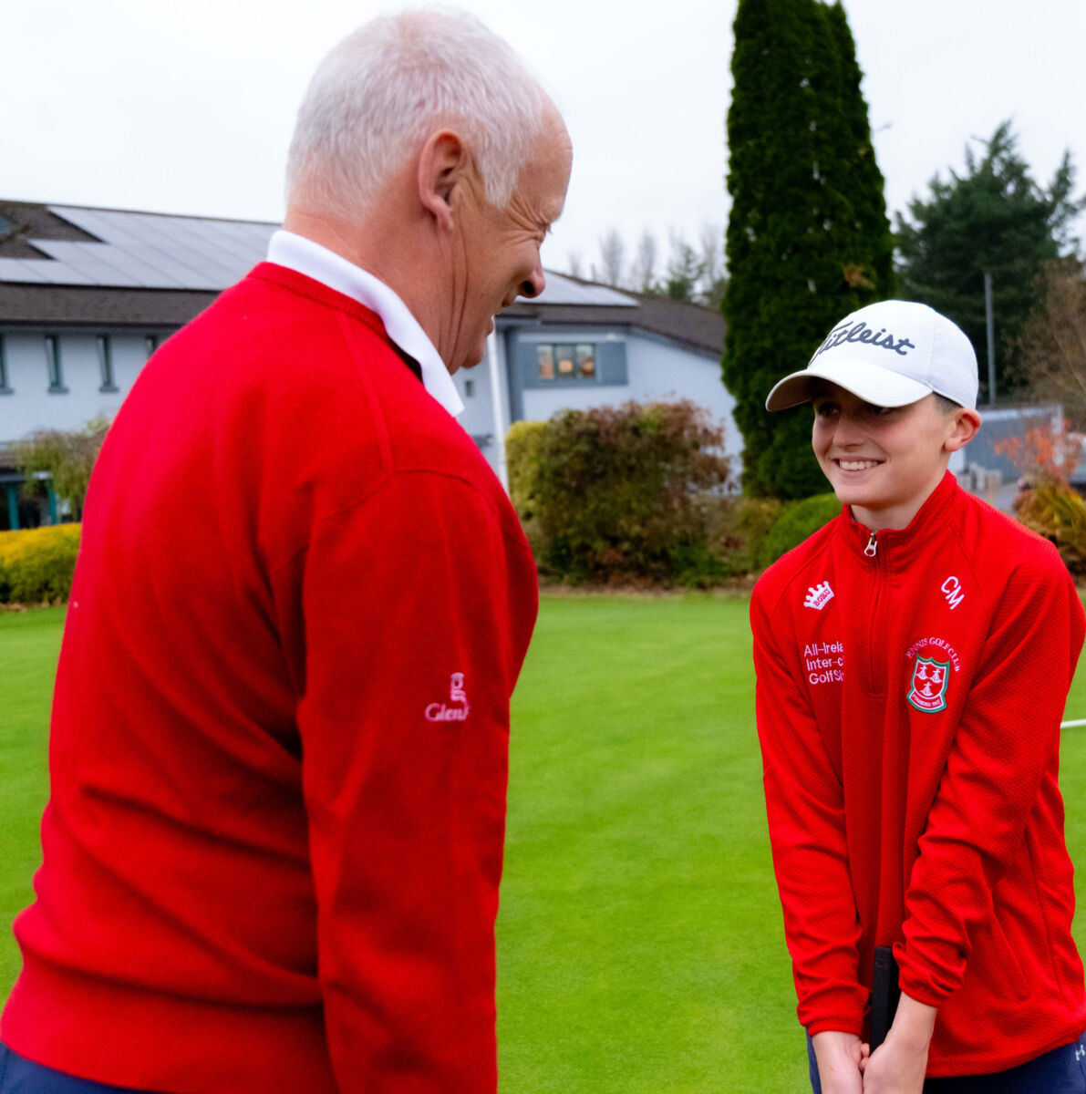 Older and younger golfer in red jackets sharing a moment on the green.