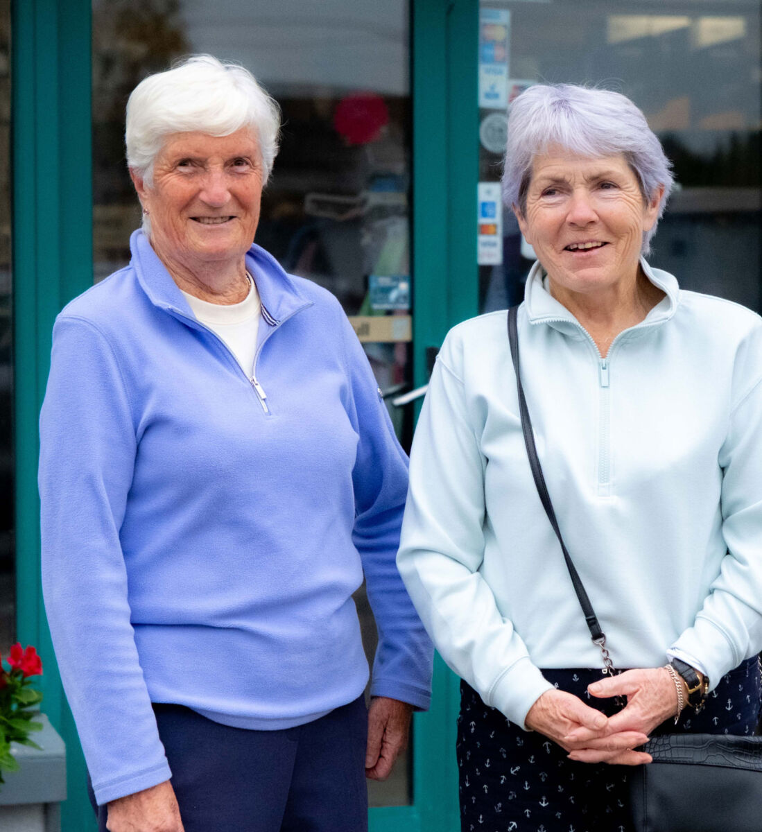 Two women smiling in front of a blue door, both wearing light jackets, one with a crossbody bag.