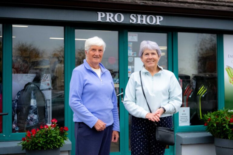 Two women smiling in front of a golf pro shop entrance with displayed merchandise.