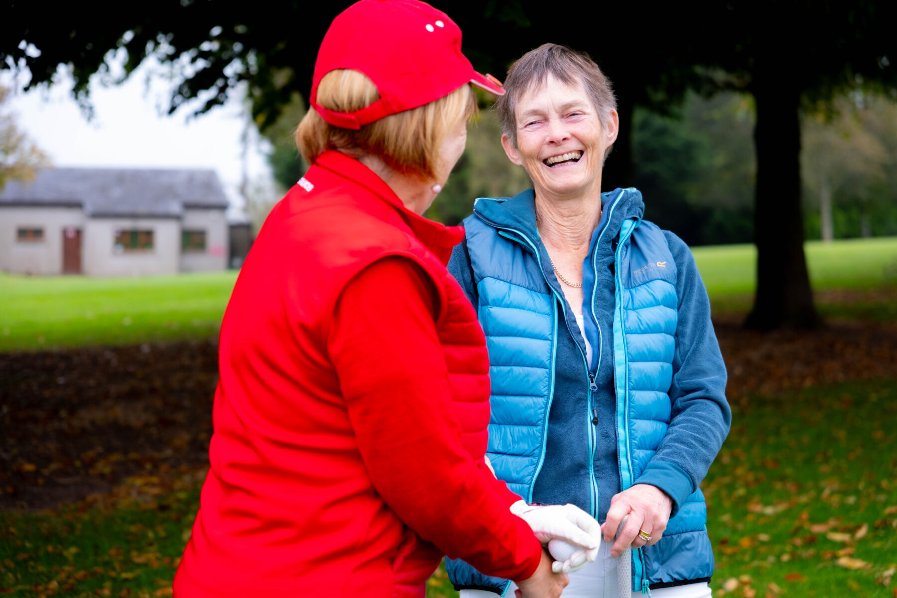 Two women smiling and chatting outdoors in colorful jackets, enjoying a pleasant day together in a park setting.