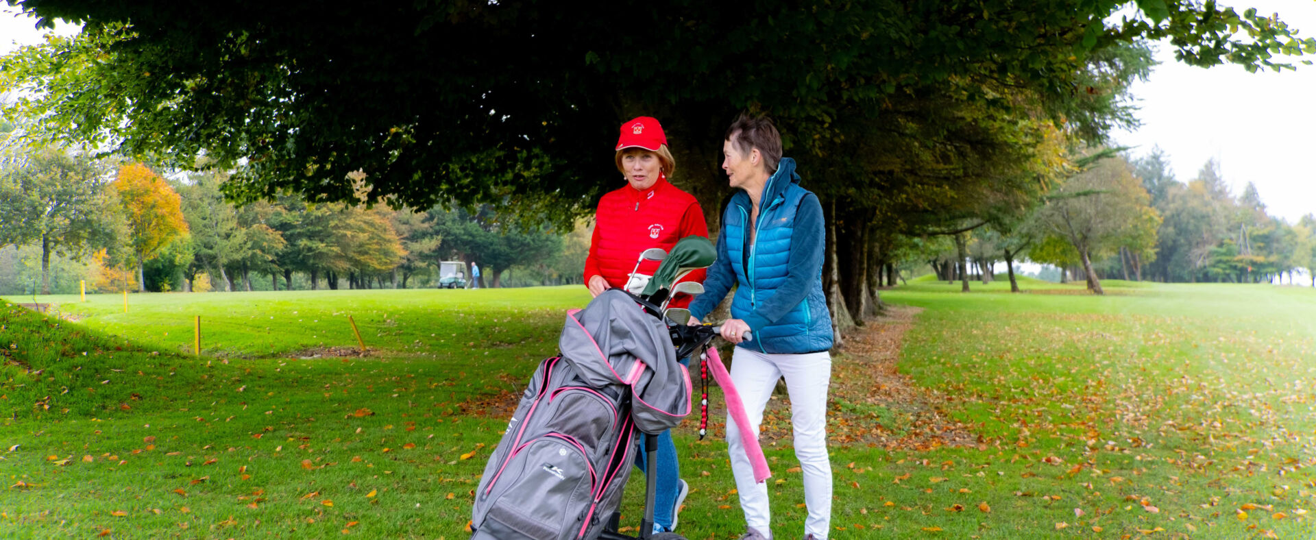 Two women walking with a golf cart on a lush green course, surrounded by autumn trees.
