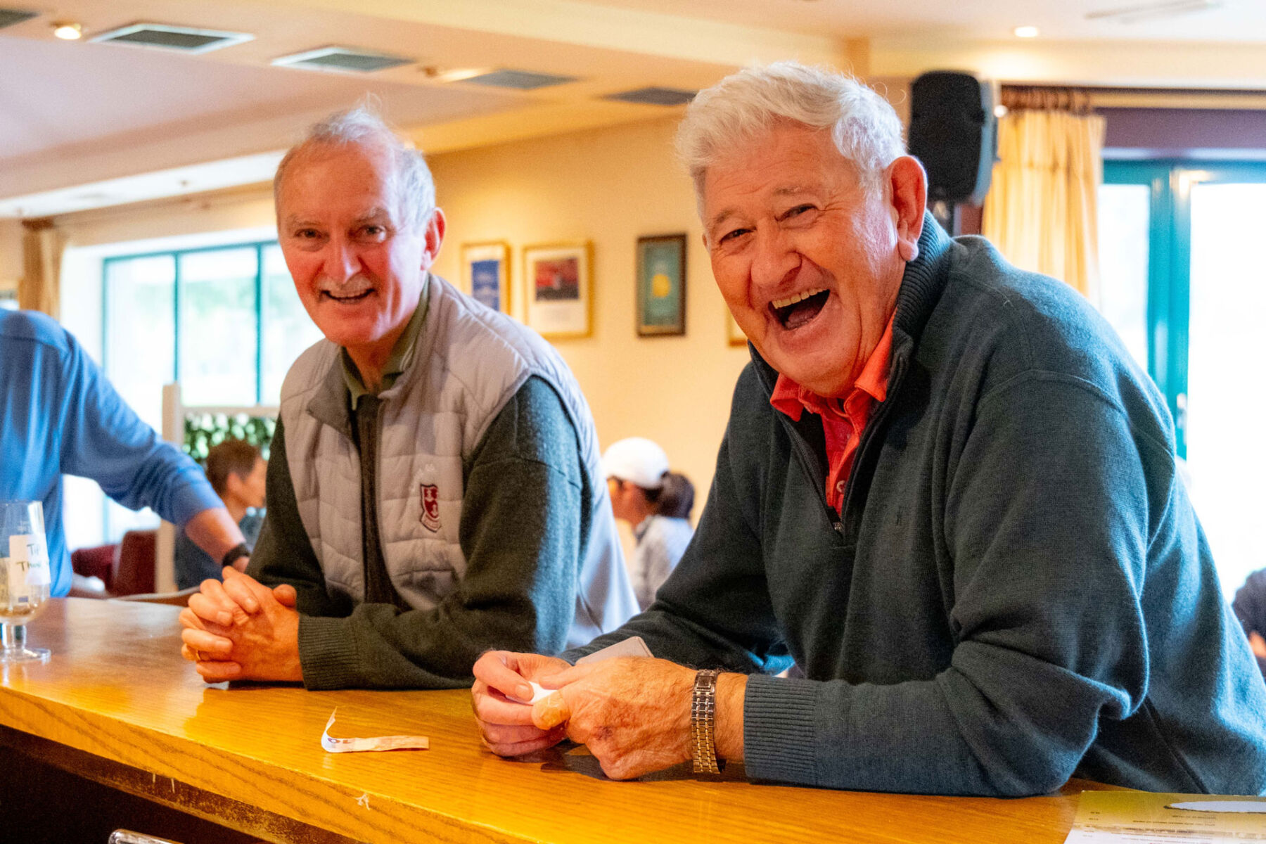 Two elderly men smiling and enjoying a conversation at a bar in a cozy, well-lit setting.