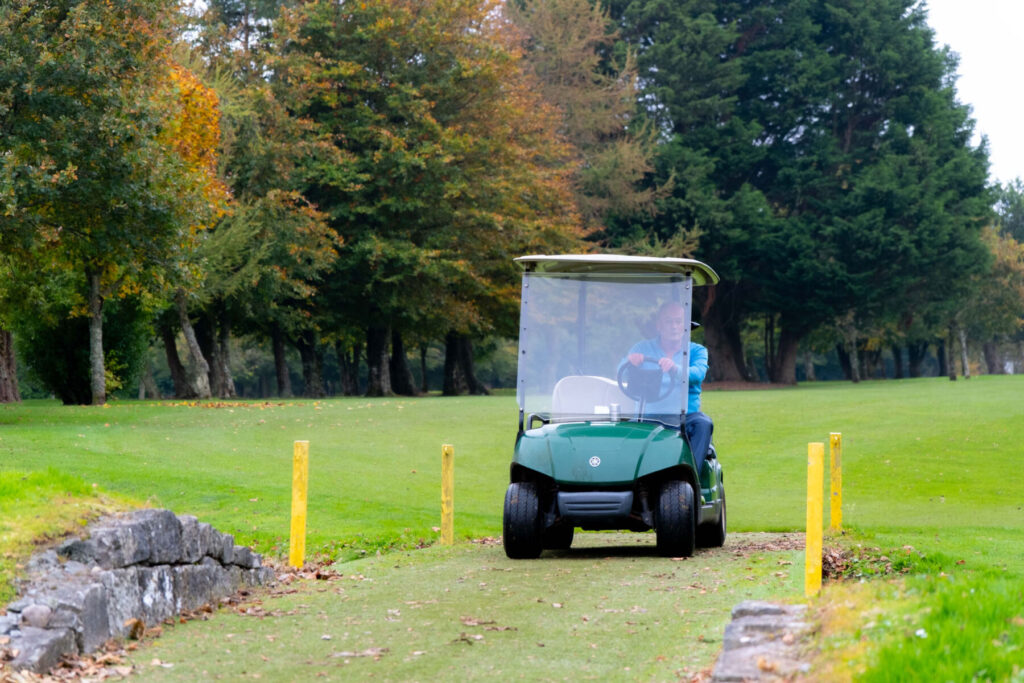 Golfer driving a green golf cart on a lush course, surrounded by vibrant trees and autumn leaves.