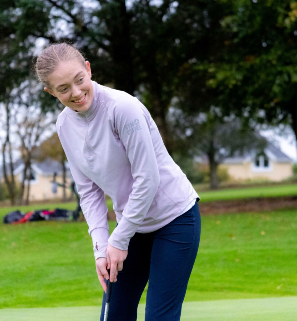 Young woman in golf attire preparing to putt on a green, with trees and houses in the background.