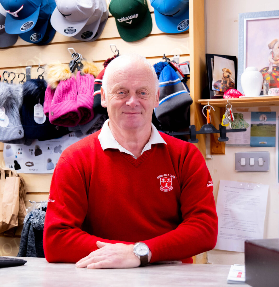 Man in red golf club sweater standing at counter, surrounded by colorful hats on display.