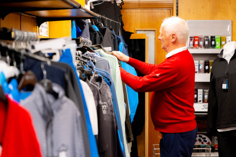 Man in red sweater shopping for clothes in a store, browsing through various jackets on a clothing rack.