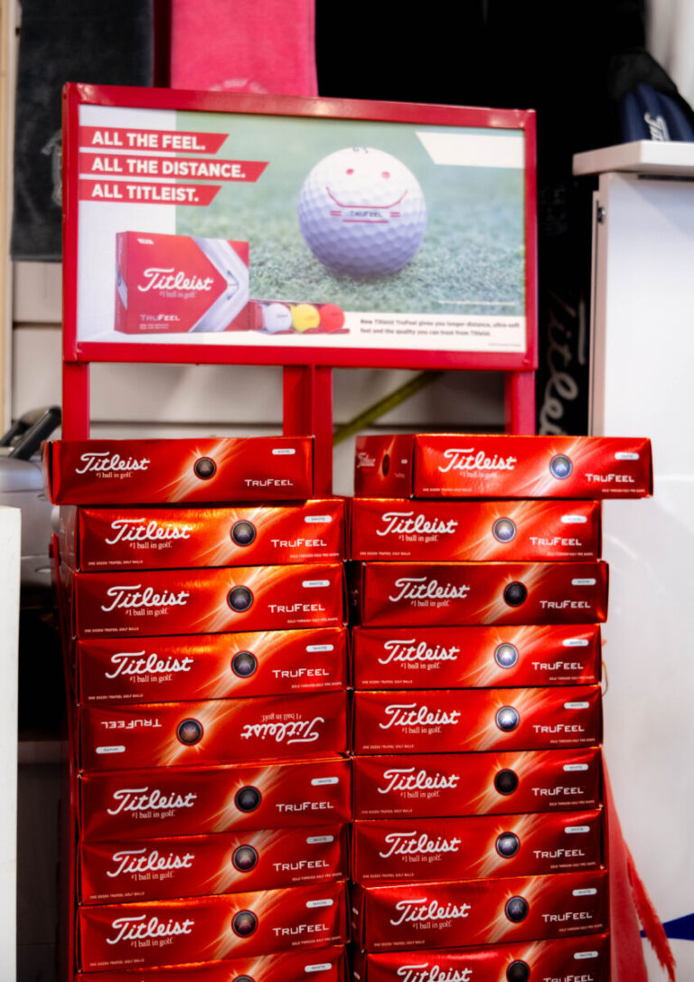 Titleist TruFeel golf ball boxes stacked with promotional sign displaying a golf ball on grass.