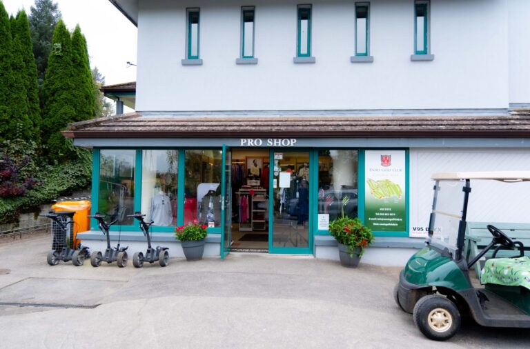 Golf pro shop entrance with golfing gear, trolleys, and cart outside, surrounded by greenery and potted plants.