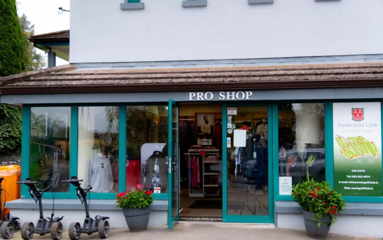 Pro shop entrance with golf apparel and equipment display surrounded by greenery and parked scooters.