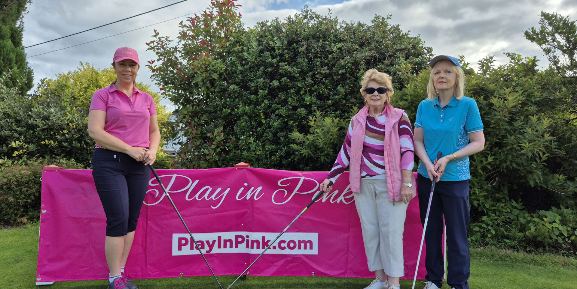 Three women with golf clubs stand beside a Play in Pink banner on a golf course.