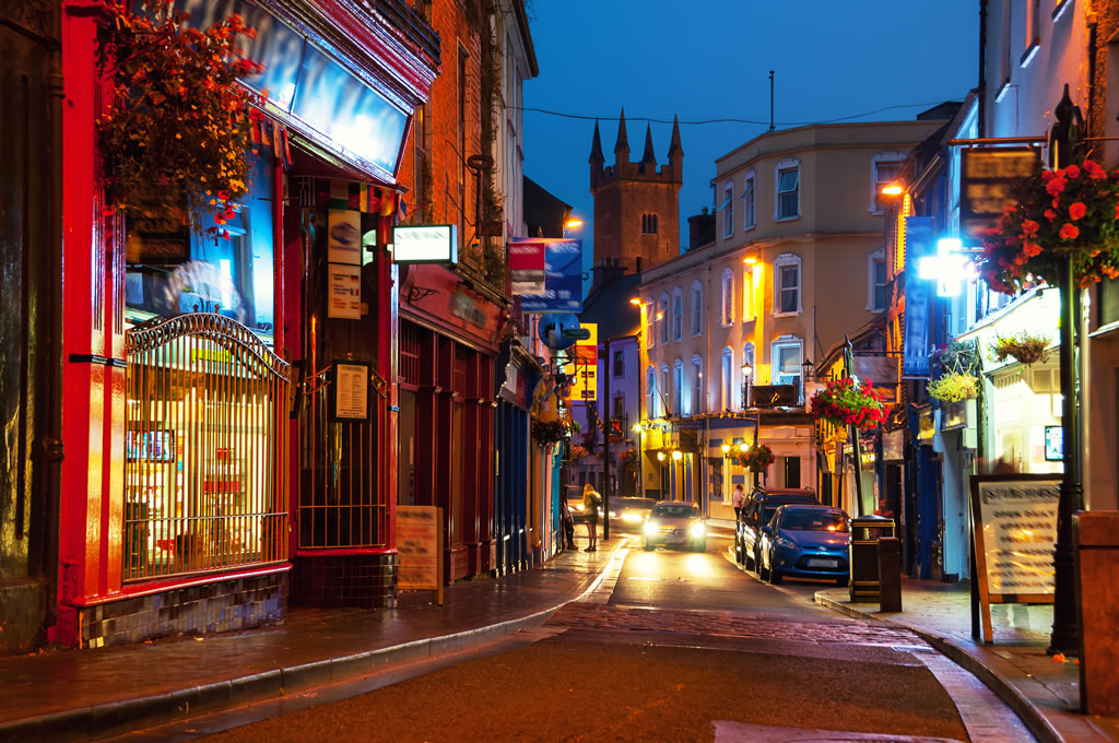 Vibrant city street at dusk with illuminated shops and a church tower in the background, decorated with hanging flowers.