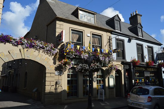 Charming pub exterior with colorful flower baskets in a sunny street setting, featuring quaint architecture and parked cars.