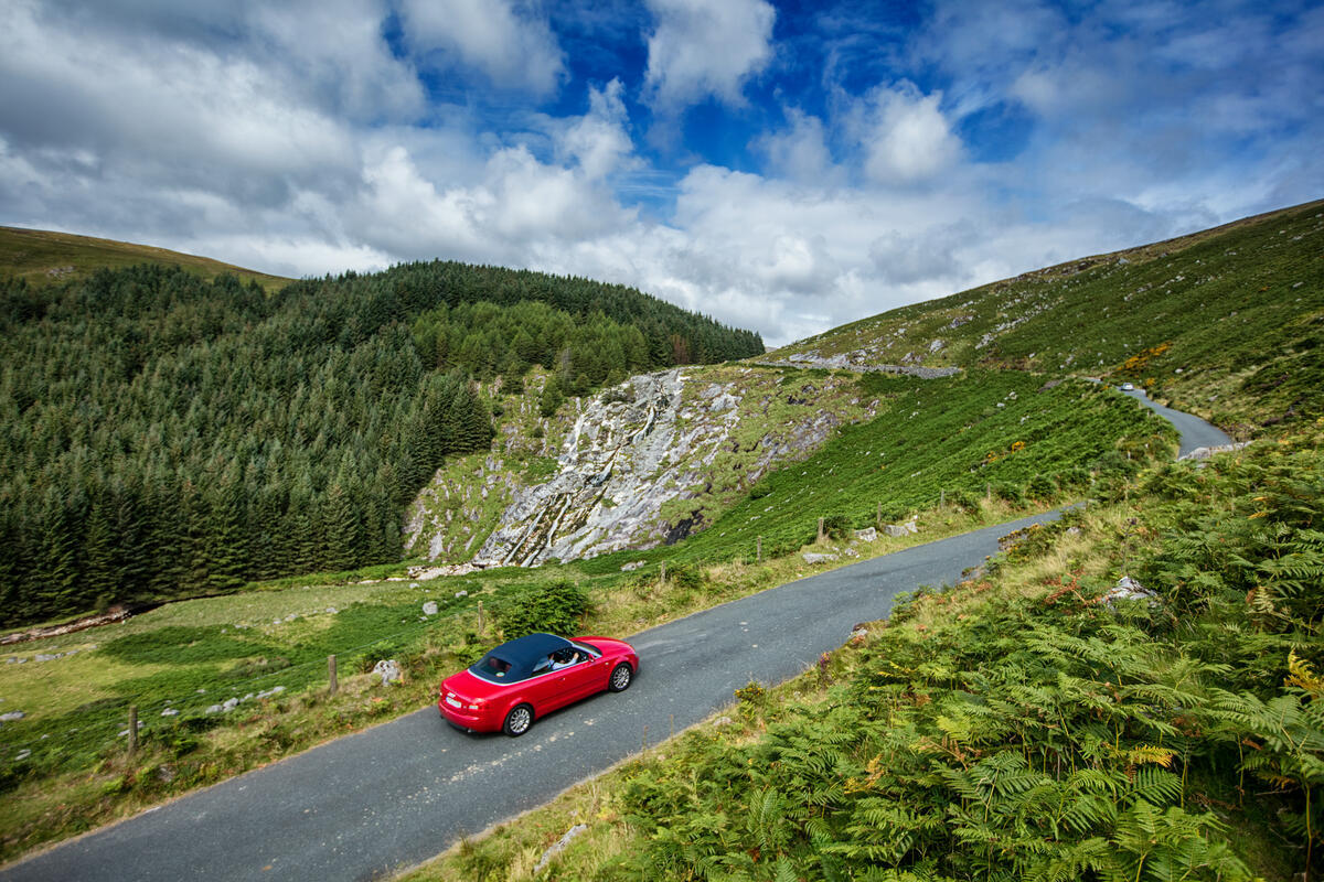A red convertible driving through scenic green hills under a cloudy blue sky.