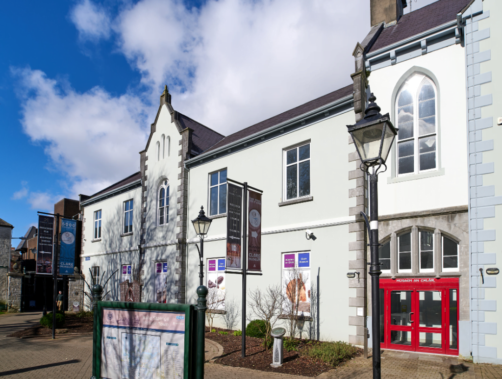 White stone building with arched windows under a blue sky, featuring a red entrance and banners on the facade.