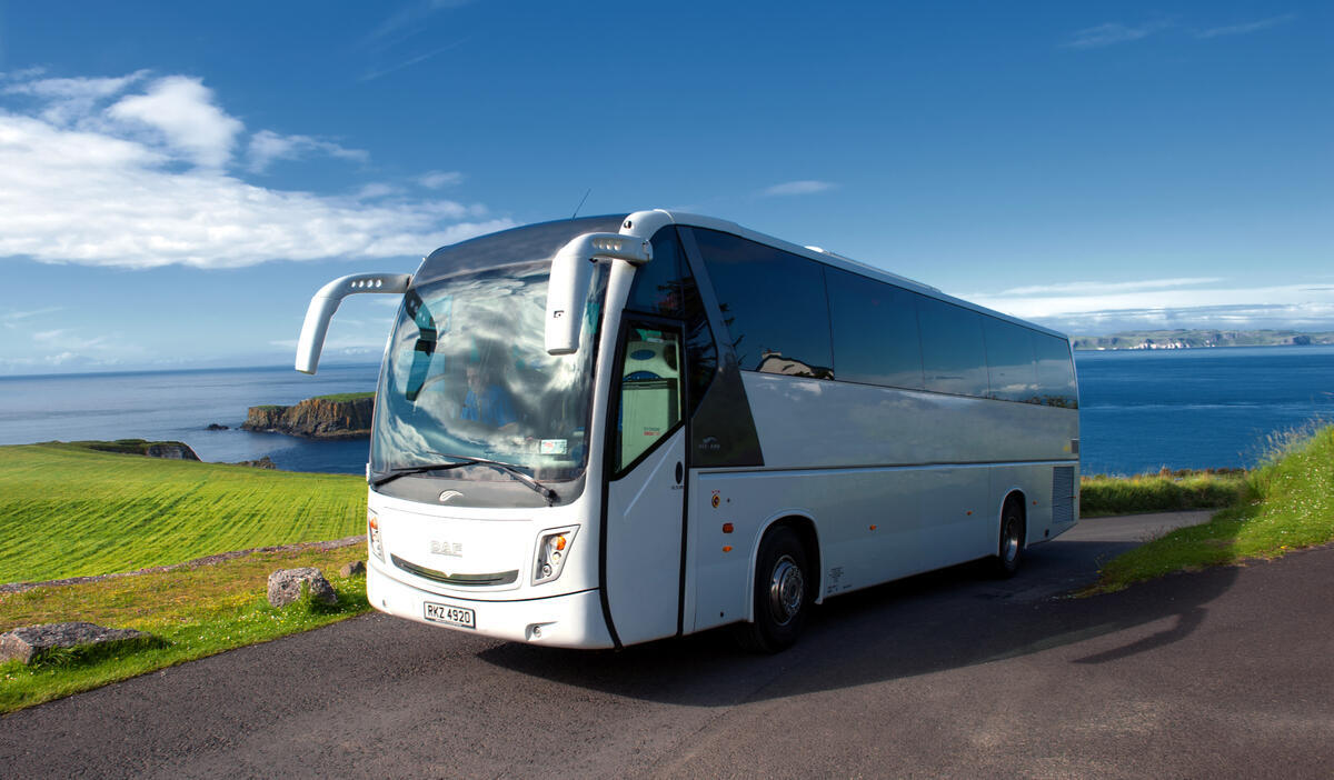 White tour bus on a scenic coastal road by the ocean under a clear blue sky.
