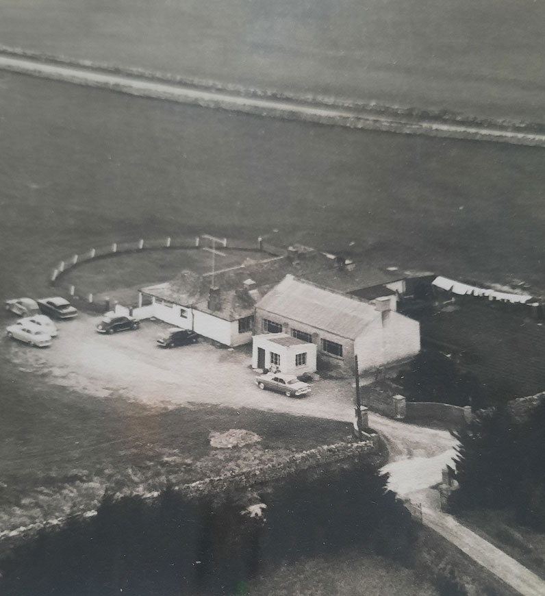 Aerial view of rural house and cars in 1960s setting, surrounded by fields and a curved driveway.