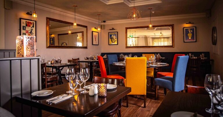 Cozy restaurant interior with colorful chairs, set tables, and warm lighting reflecting off large mirrors on the walls.