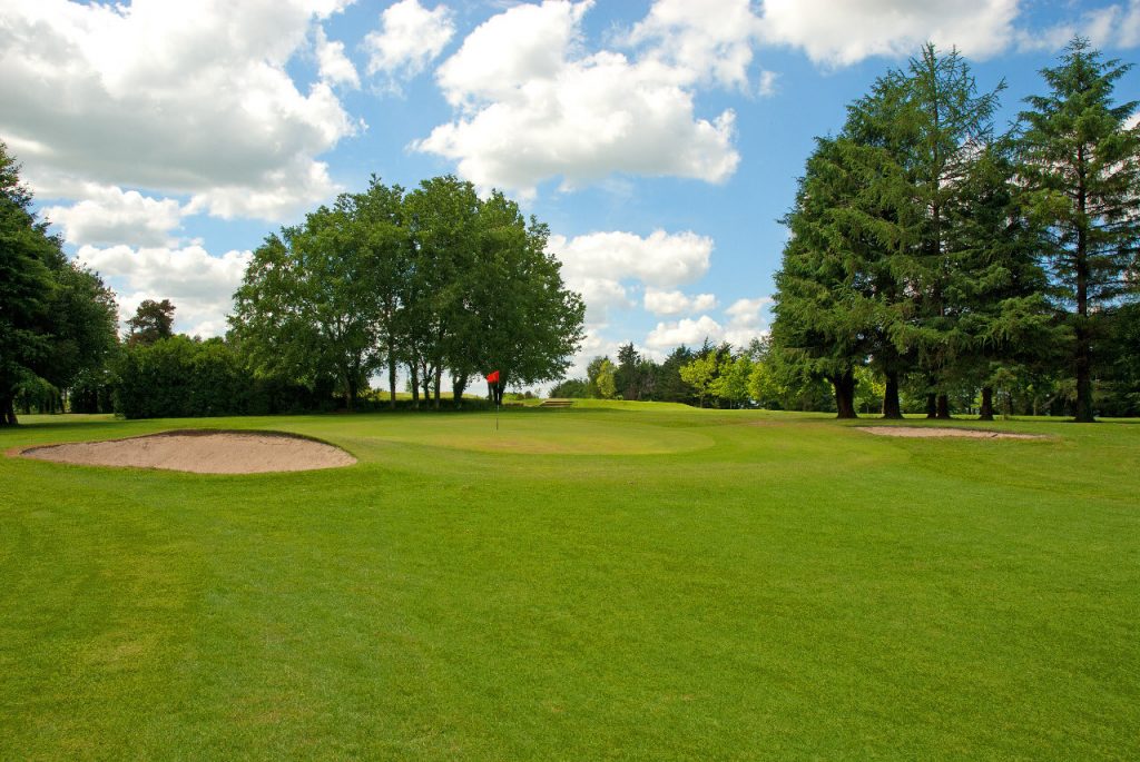 Golf course with sand traps, green grass, and a flag under a blue sky with clouds. Perfect day for golfing.