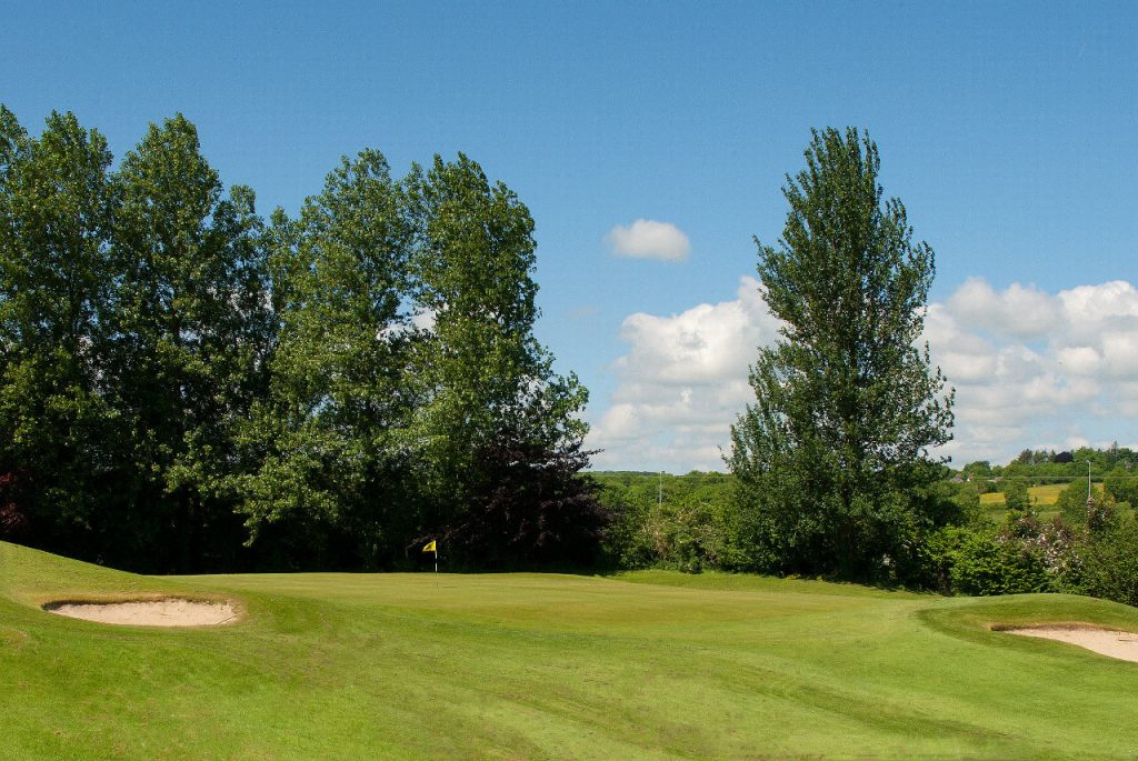 Golf course green with sand bunkers and trees under a blue sky on a sunny day.
