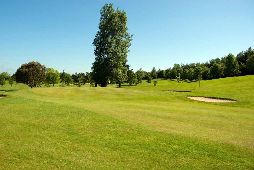 Wide golf course landscape with lush green fairway, sand bunkers, and trees under a clear blue sky.