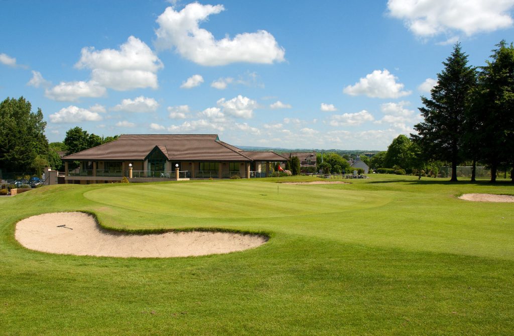 Golf course with clubhouse, green grass, and sand bunkers under a blue sky with clouds.