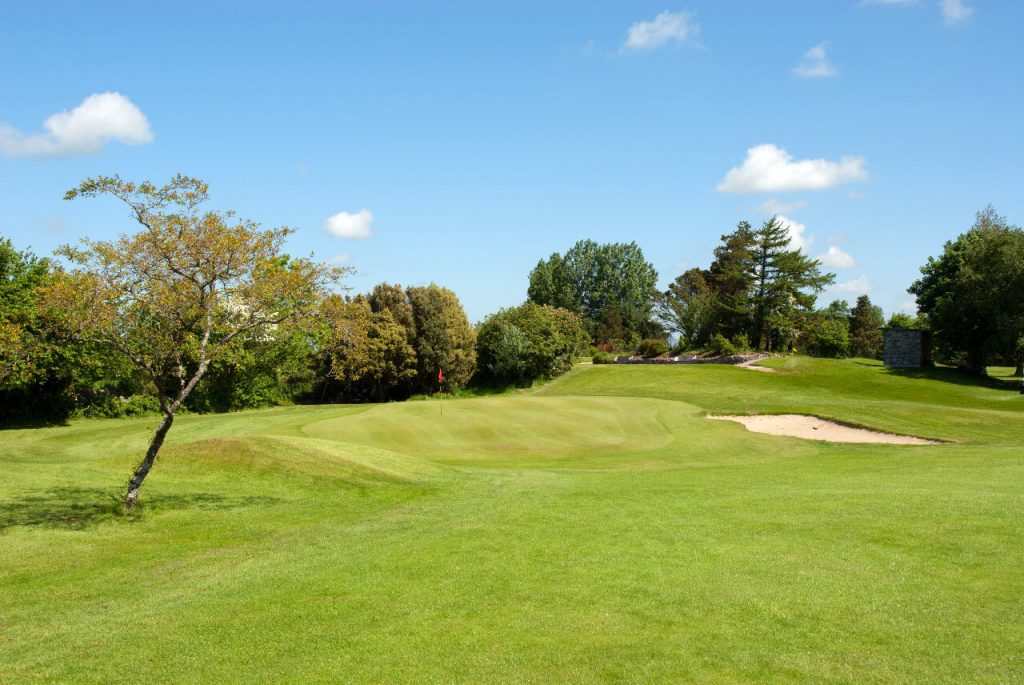 Scenic golf course with lush green fairway, sand bunker, and vibrant trees under a blue sky with clouds.