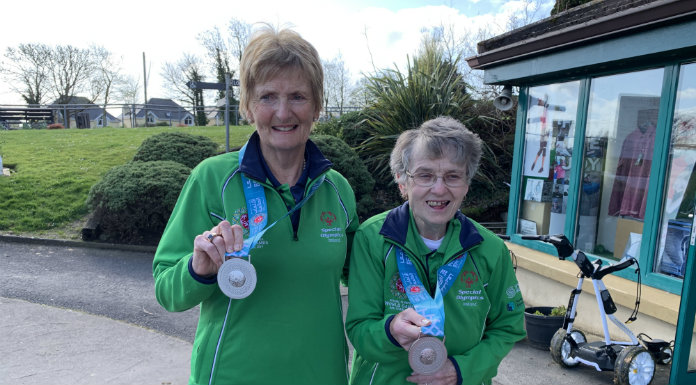Two athletes proudly display their medals outdoors, wearing green jackets and smiling.