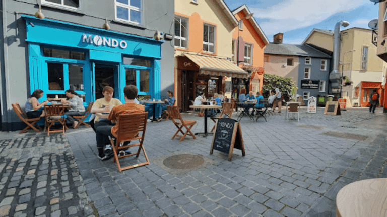 Outdoor cafe scene with colorful buildings and patrons enjoying meals on a cobblestone street.