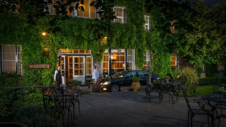 Ivy-covered hotel entrance with a parked car and outdoor dining area illuminated at night.