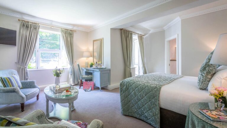 Elegant hotel room with cozy seating area, large bed, soft lighting, and garden view through large windows.