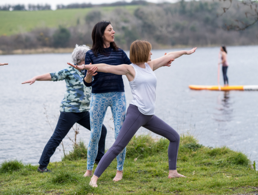 Women practicing yoga outdoors by a lake, with a paddleboarder in the background.
