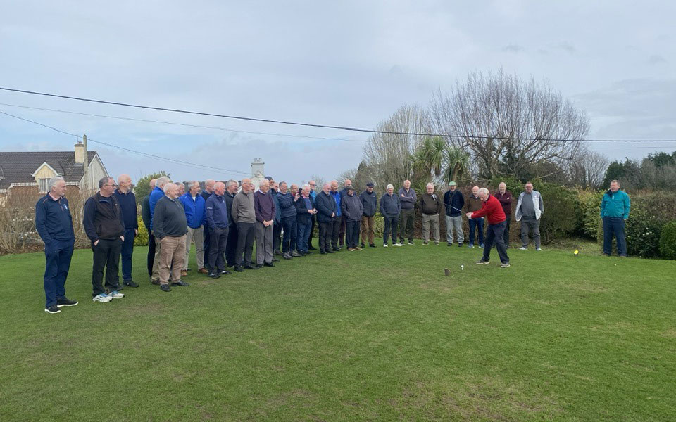 Group of seniors watching a man in red sweater preparing to tee off on a golf course on a cloudy day.