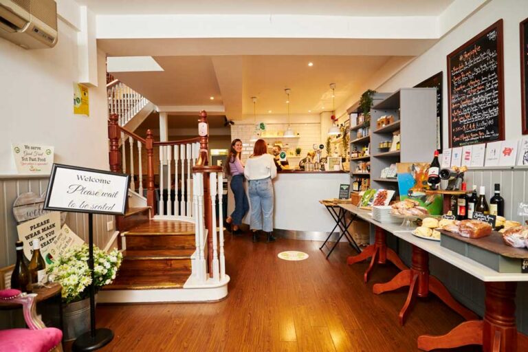 Cozy cafe interior with customers at the counter, baked goods displayed on a table, and welcoming signs at the entrance.