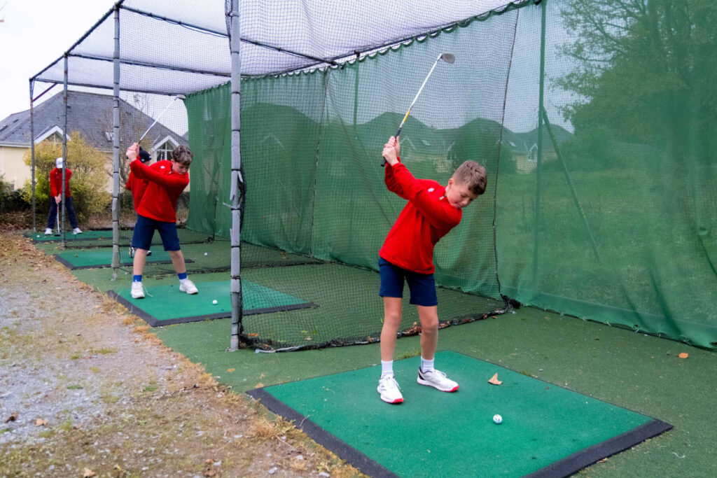 Kids practicing golf swings in a netted range wearing red tops and shorts, focus on technique improvement.