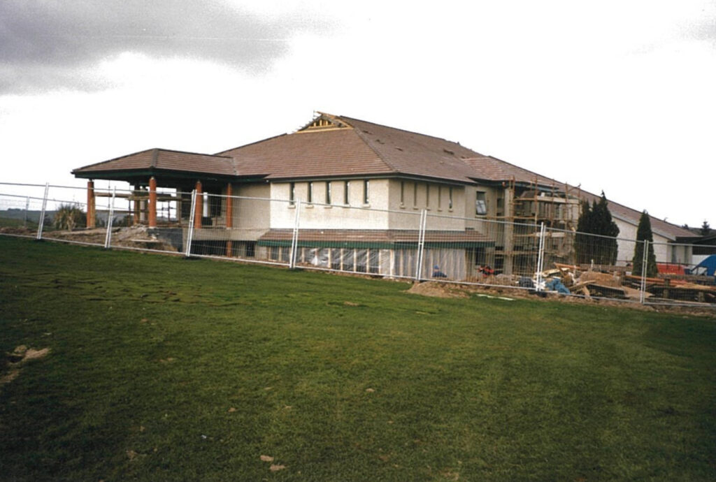 Under-construction building with fenced perimeter and green lawn in foreground.