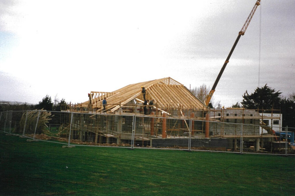 Construction site with workers building a wooden roof frame supported by a crane, enclosed by fencing.