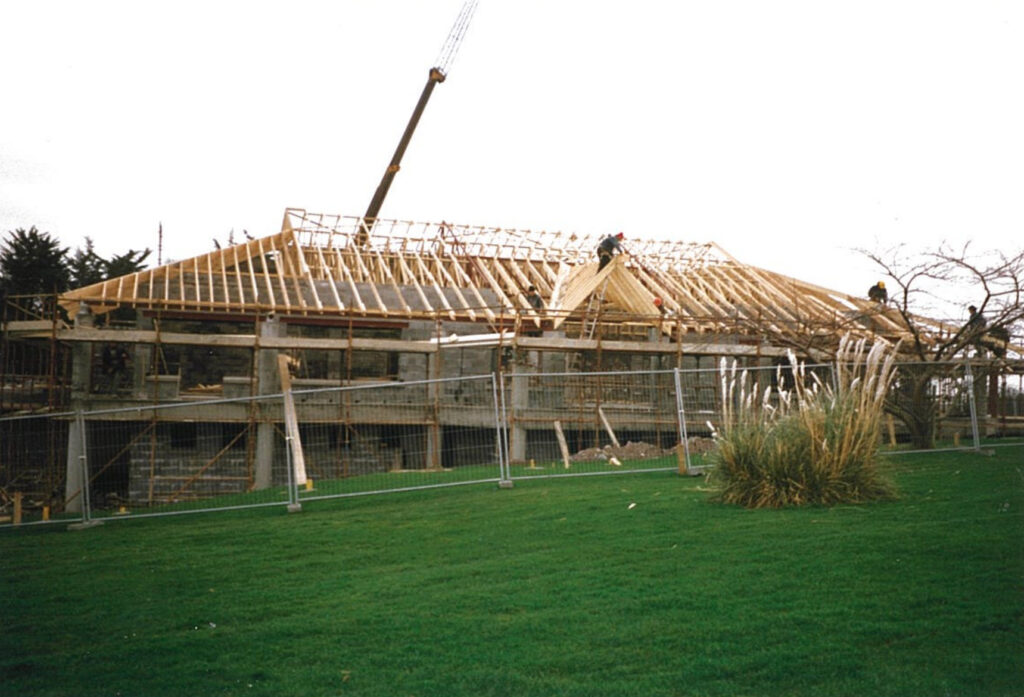 Building under construction with wooden roof trusses and crane, surrounded by fence and green lawn.