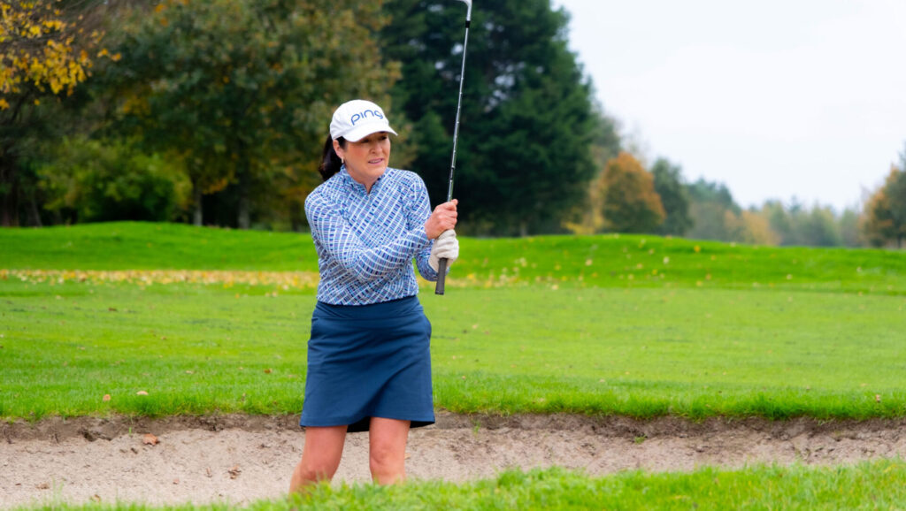 Woman playing golf in blue attire at a lush green golf course, swinging a club near a sand bunker.