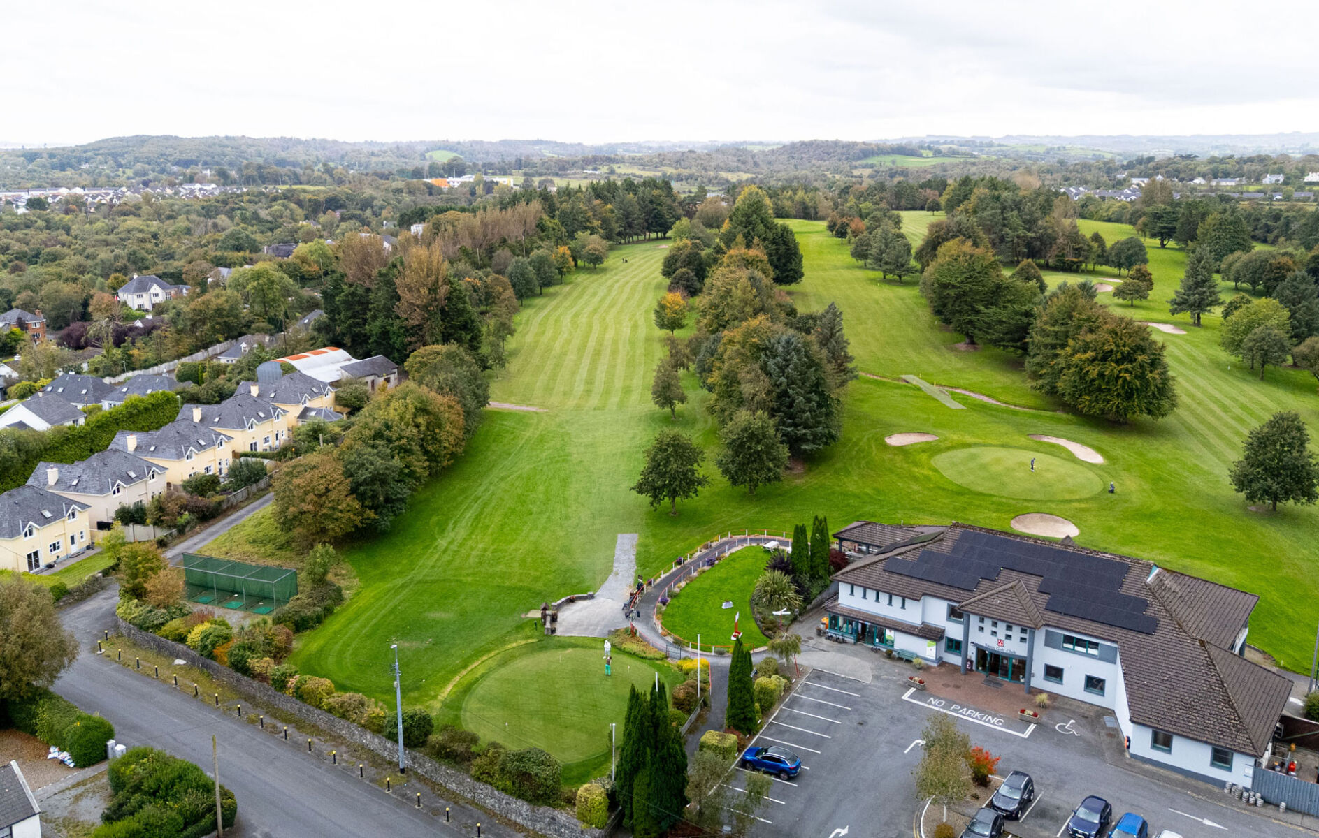 Aerial view of a golf course with lush greens, clubhouse, and nearby residential area under a cloudy sky.