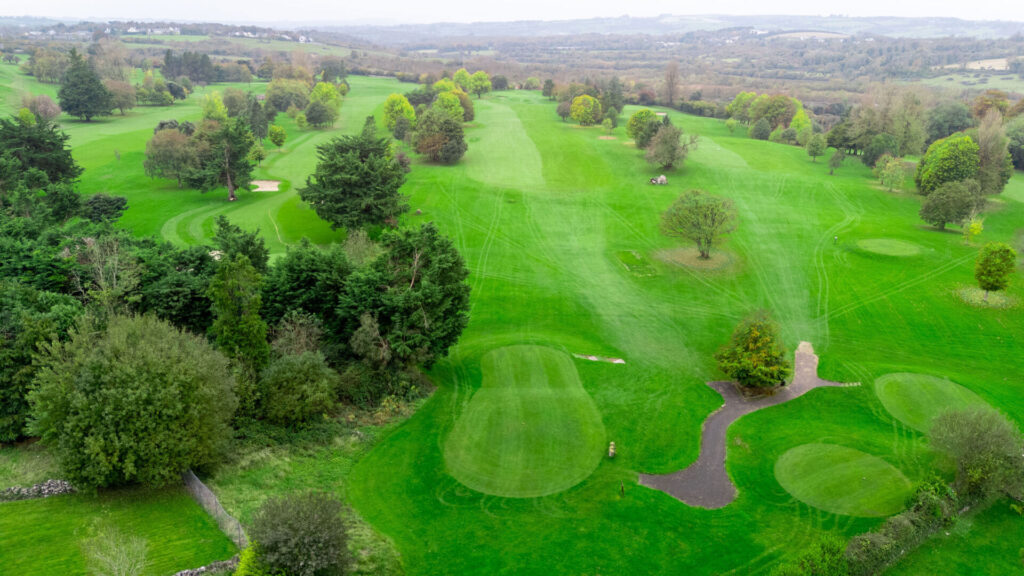 Aerial view of lush green golf course with trees, fairways, and bunkers under a cloudy sky.
