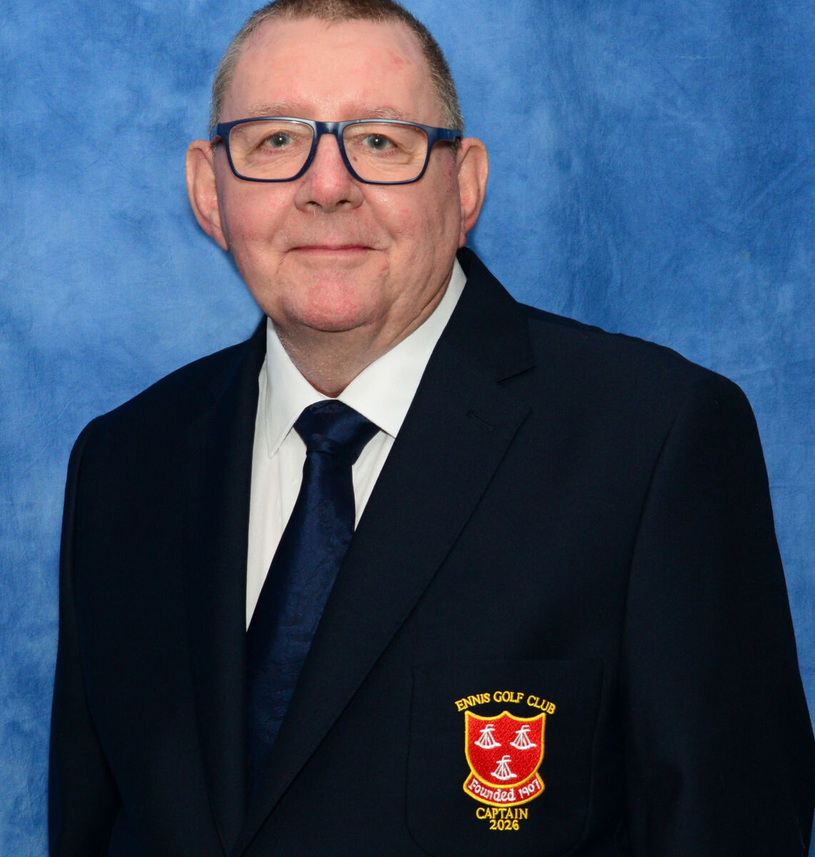 Man in a suit with glasses and Ennis Golf Club badge, stands against a blue background.
