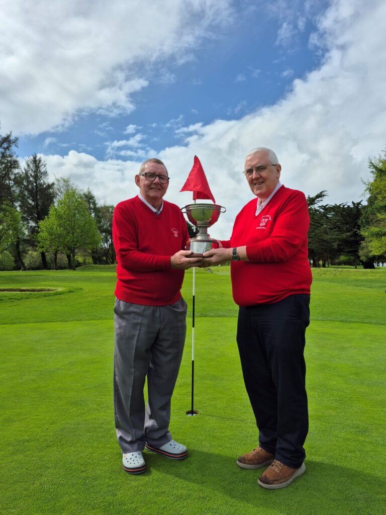 Two men in red sweaters hold a trophy on a golf course with a clear sky in the background.