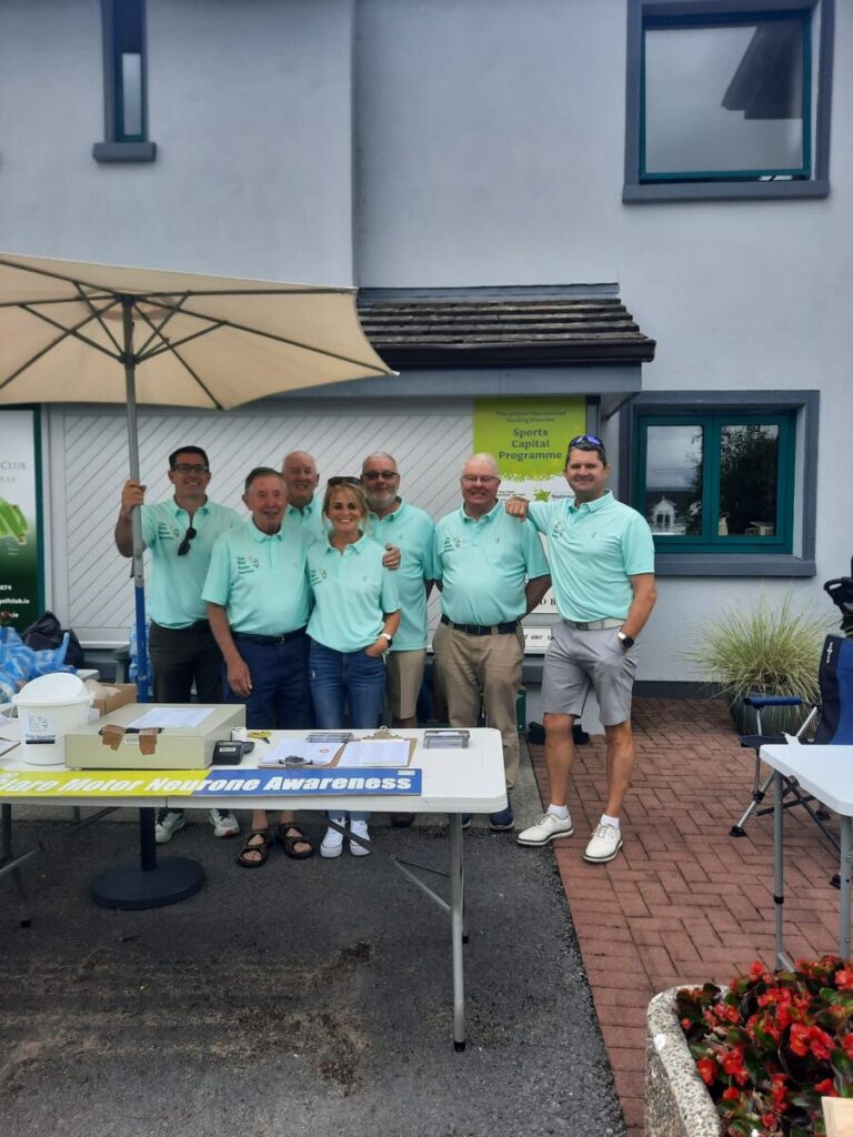 Group of volunteers in light blue shirts at a motor neurone awareness event with an informational table and umbrella.