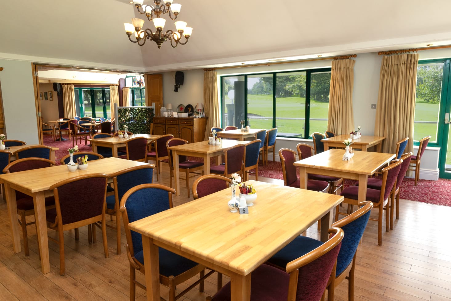 Elegant dining room with wooden tables and chairs, large windows overlooking a green landscape, and a chandelier ceiling.