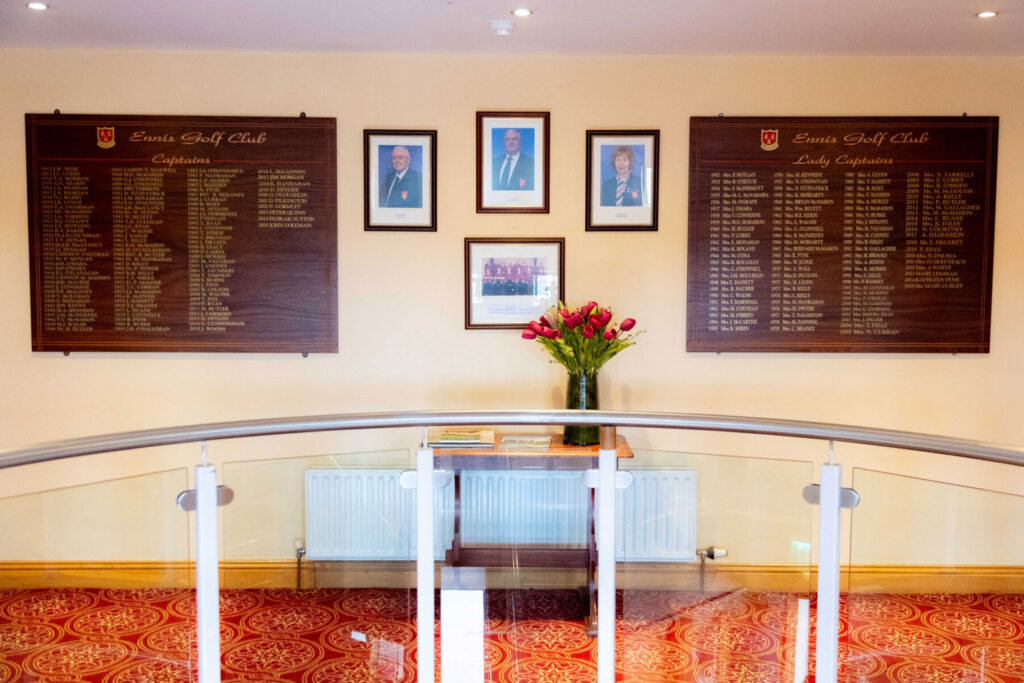 Ennis Golf Club captains wall with photos and flower vase on a wooden table in a decorated hallway.